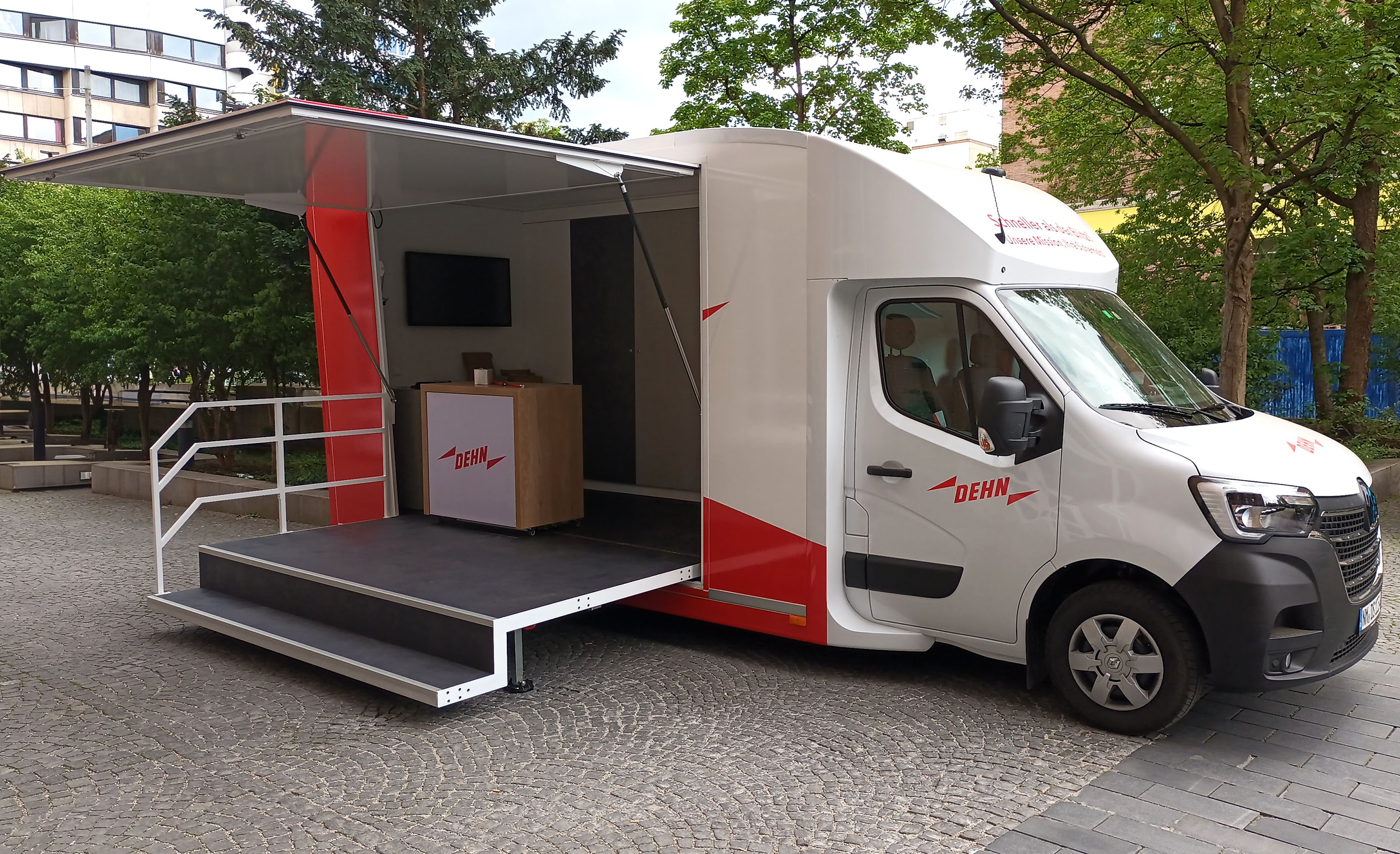 Das DEHN Blitzschutzmobil steht geöffnet auf einem gepflasterten Platz mit Bäumen im Hintergrund