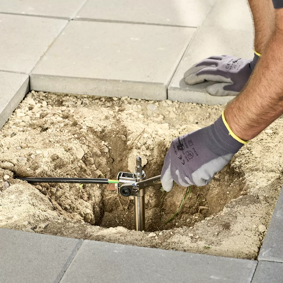 Montage einer Erdungsanlage: Eine Person mit Arbeitshandschuhen befestigt eine Erdungsstange in einem ausgehobenen Erdloch zwischen Betonplatten. Die Stange wird mit einem Schraubenschlüssel und einer Verbindungsklemme justiert, um eine sichere elektrisch