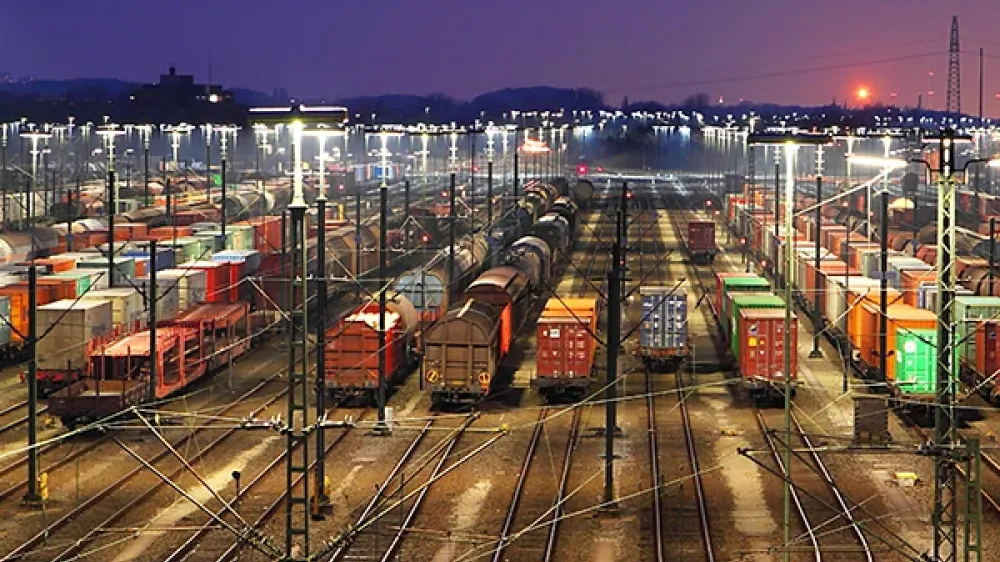 Bahnhofgelände mit vielen Gleisen, Weichen und Stromleitungen vor der Silhouette einer Stadt.