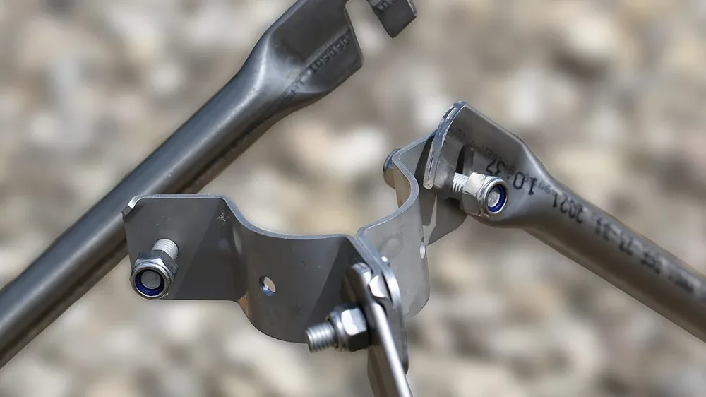Close-up of metal rods connected by a clamp with bolts and nuts, showing open-ended joints on a blurred gravel background.