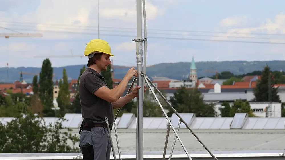 Person wearing a yellow helmet adjusts a metal mast with support rods on a gravel rooftop, buildings visible in the background.