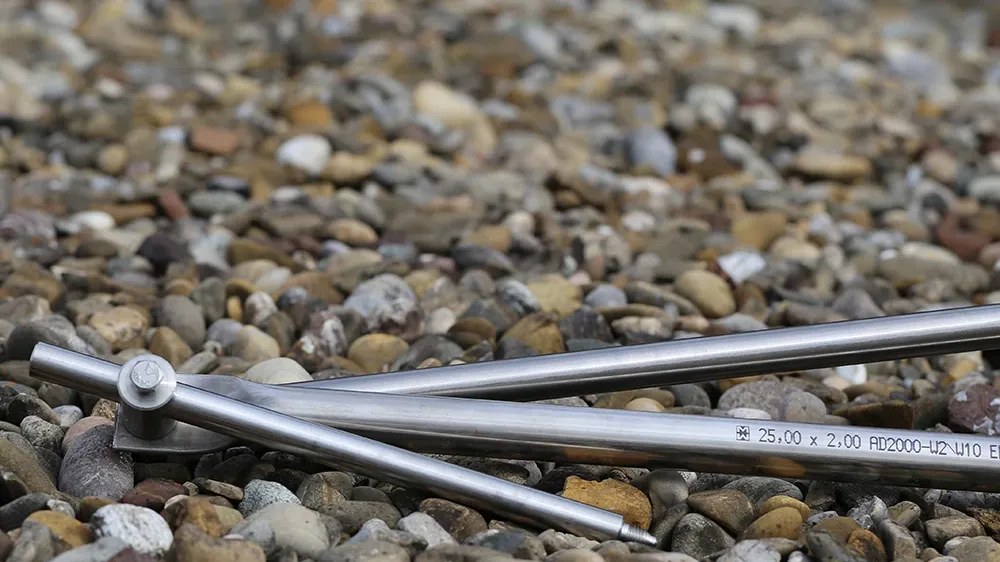 Metal rods with pointed ends lying on gravel, hinge joint visible, printed text “25,00 x 2,00 AD2000-UZ V10 E” on one rod.