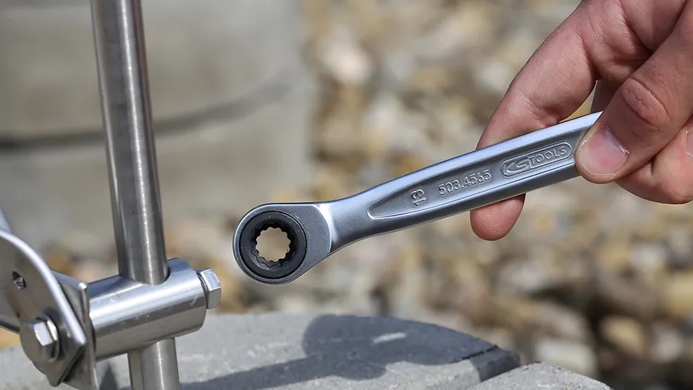 Hand using a ratcheting wrench to tighten a clamp on a vertical metal rod fixed into a concrete block.