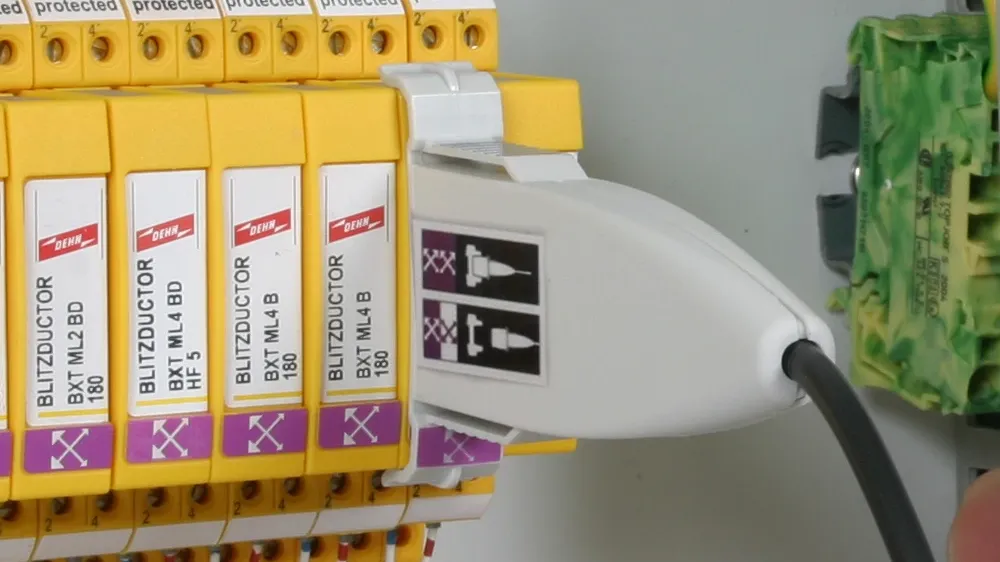 White test adapter plugged into a yellow surge protection module inside an electrical control cabinet with connected wires.