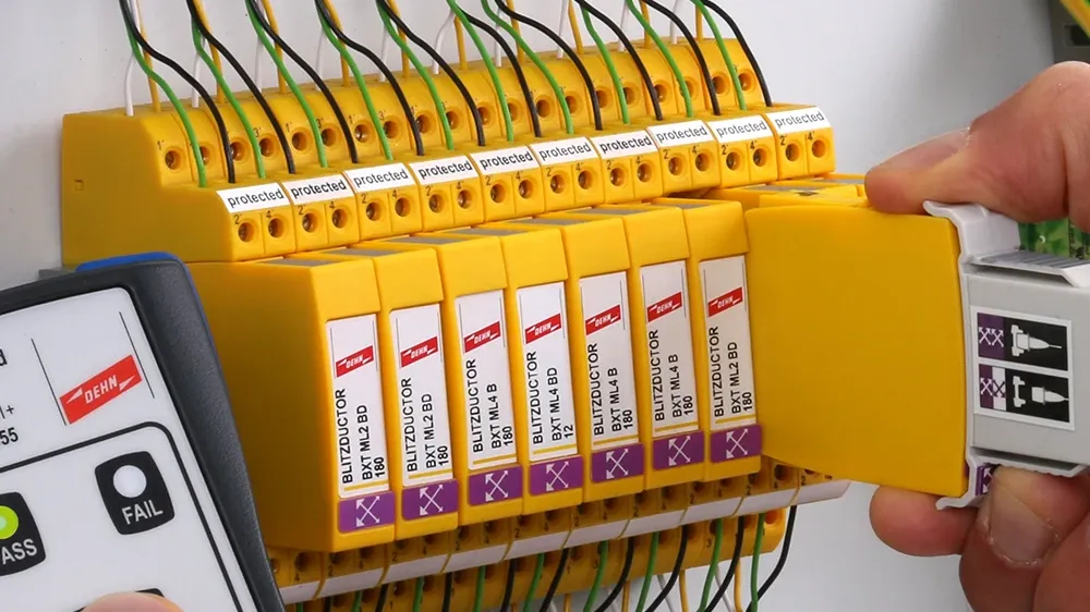 Hand holding a measuring device next to a row of yellow surge protection modules while another hand holds a removed module with test adapter.