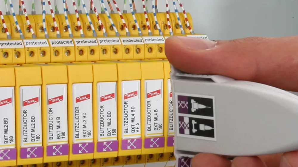 Hand holding a white test adapter next to yellow surge protection modules inside a control cabinet with connected wires.