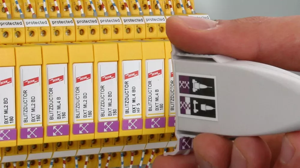 Hand holding a white test adapter next to yellow surge protection modules inside a control cabinet with connected wires.