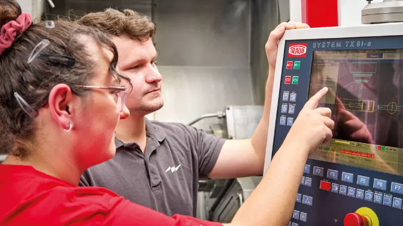 Two people operate a machining control panel with a screen showing technical diagrams.