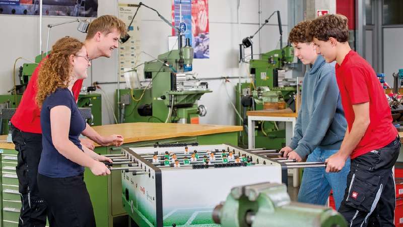 Vier junge Leute, zwei auf jeder Seite, spielen Tischfußball in einer Industriehalle. Sie lächeln und sind konzentriert, im Hintergrund sind grüne Maschinen zu sehen.