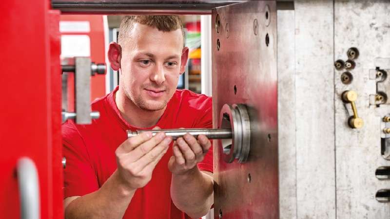 Ein Mann in einem roten Hemd prüft oder montiert ein Metallteil an einer Industriemaschine in einer Werkstatt.