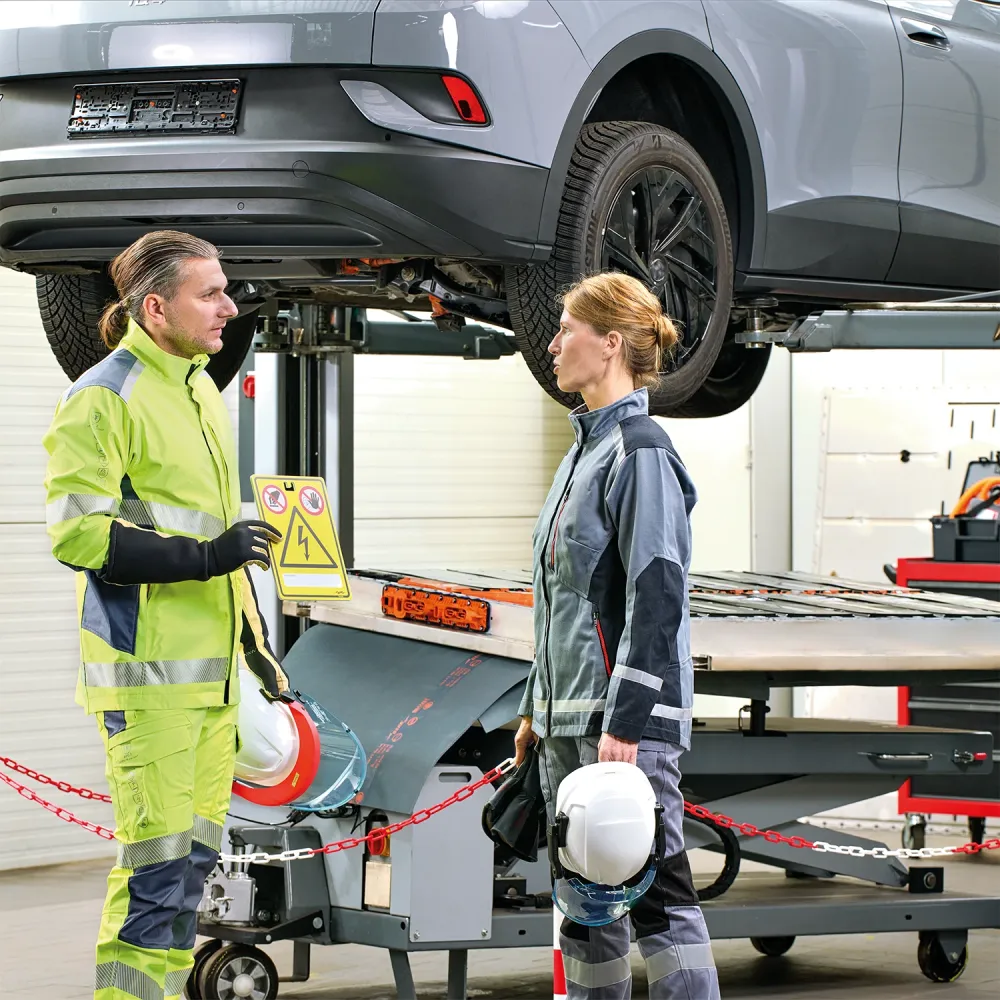 Zwei Arbeiter in Schutzkleidung diskutieren unter einem angehobenen Elektrofahrzeug. Einer hält ein Warnschild. Beide tragen Helme und Warnschutzkleidung. In der Garage sind Werkzeuge und Maschinen zu sehen.
