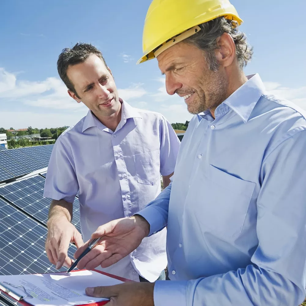 Two men are next to a ground-mounted PV system. One wears a construction helmet and a clipboard in his hand, which both people point to.
