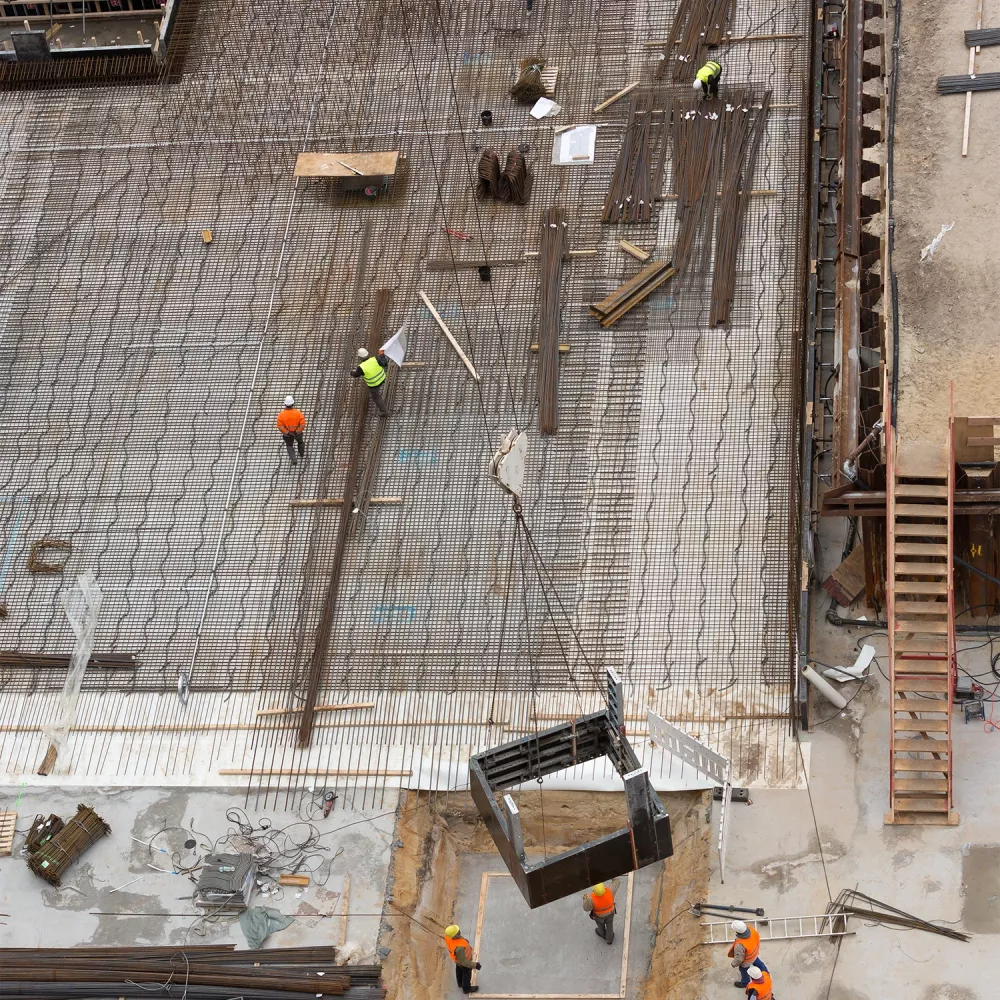 Overhead view of a construction site with two workers in safety helmets walking on a steel reinforcement grid with materials and equipment nearby.