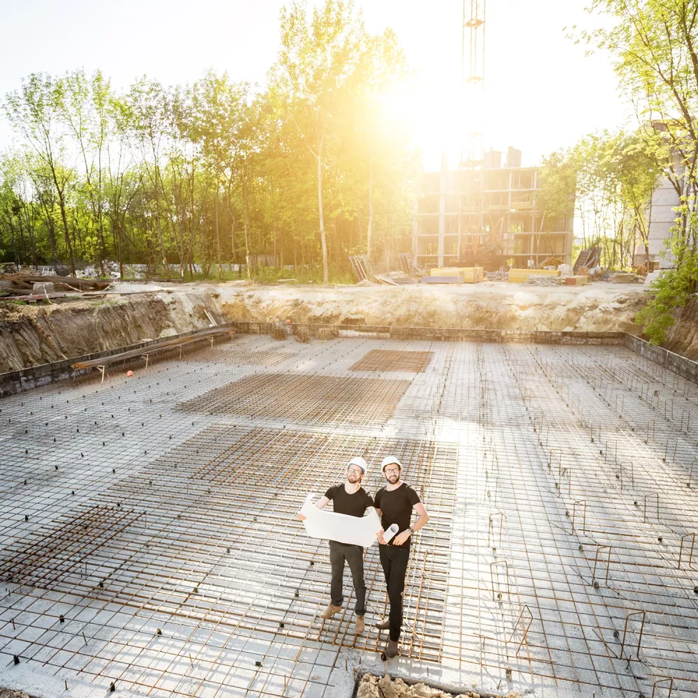 Two construction workers wearing hard hats stand on a reinforced concrete foundation slab, holding blueprints at a building site.