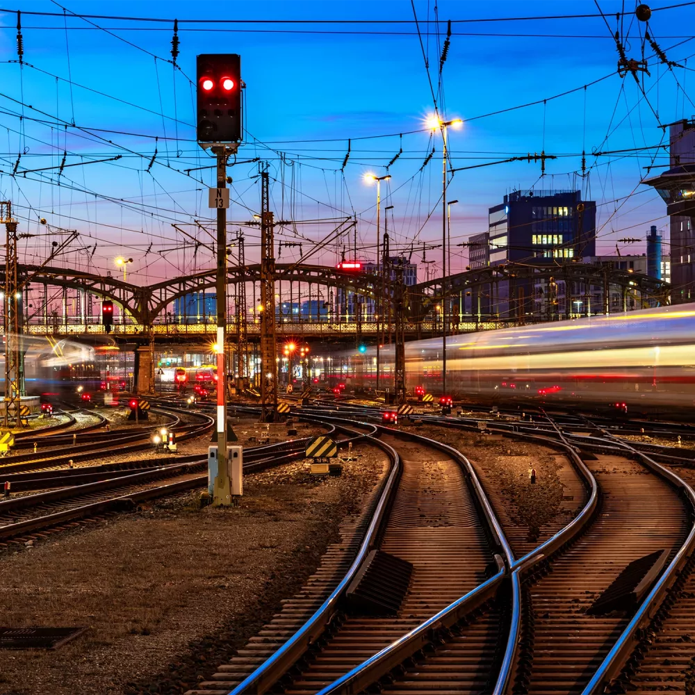 Railway junction at dusk with multiple tracks, overhead power lines, red signal lights, and blurred trains passing through an urban station area.