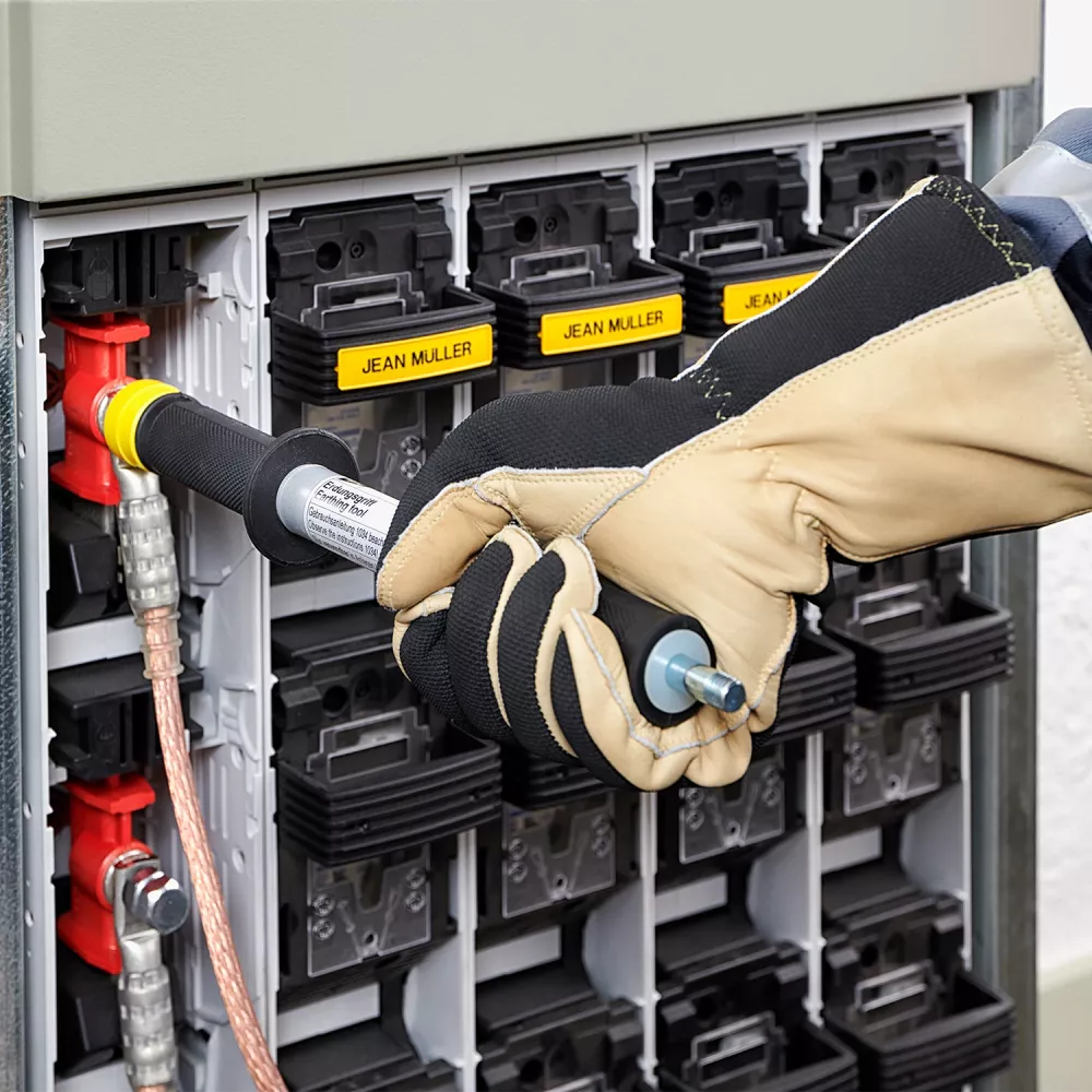 Close-up of a gloved technician connecting a grounding tool to a fuse switch in an electrical distribution cabinet with labeled modules.