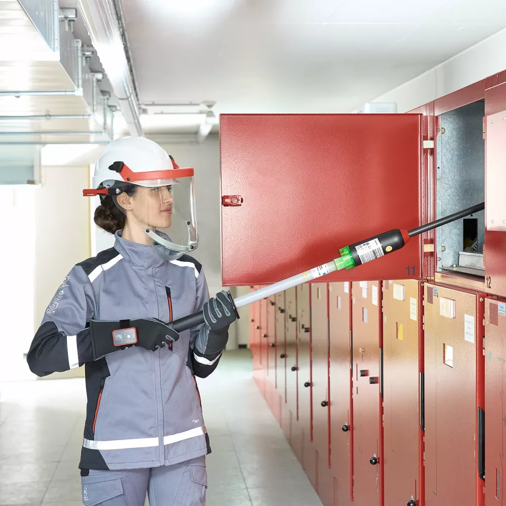 Technician wearing face shield and protective clothing uses an insulated testing pole on an open red electrical switchgear cabinet.