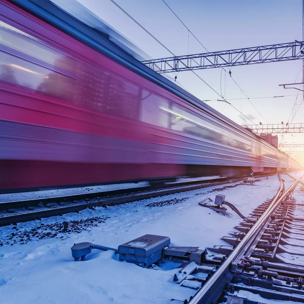 High-speed train passing a snow-covered railway junction at dusk, with blurred motion, overhead power lines, and visible switch components along the tracks.