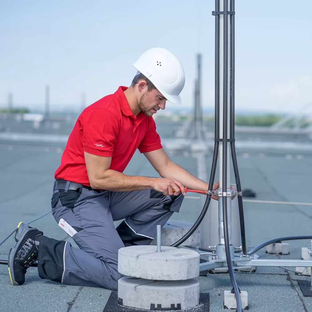 Technician in red shirt and white hard hat installing grounding cables on a rooftop lightning protection mast.