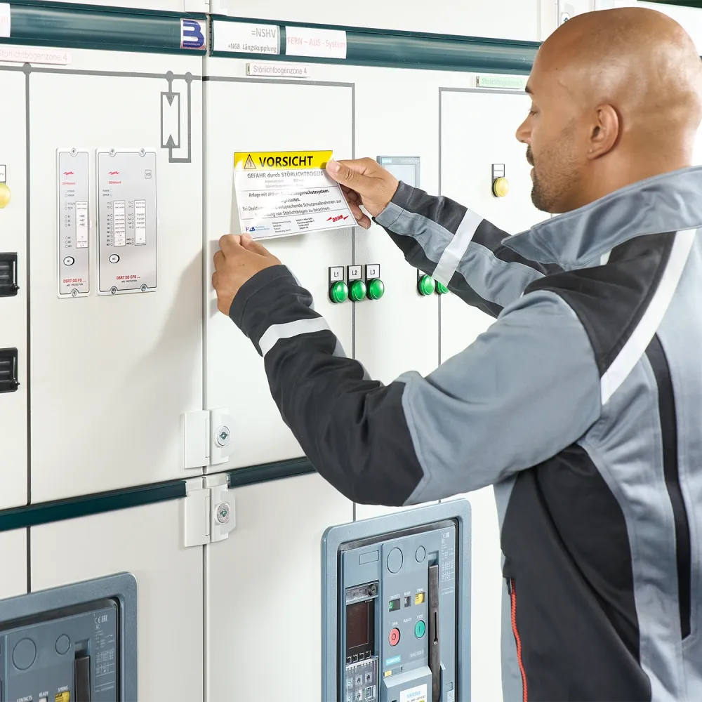 Technician attaching a warning label to an electrical control cabinet with switches, indicator lights, and monitoring devices.