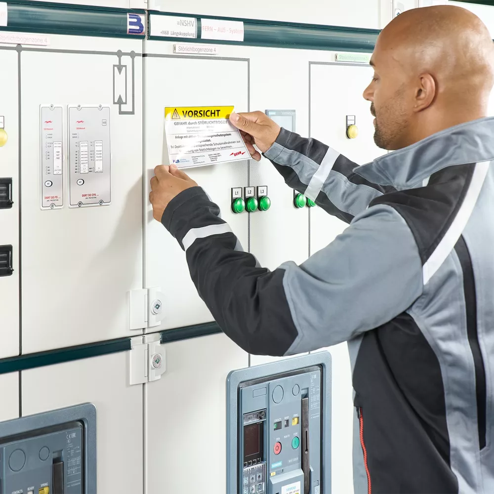 Technician attaching a warning label to an electrical control cabinet with switches, indicator lights, and monitoring devices.