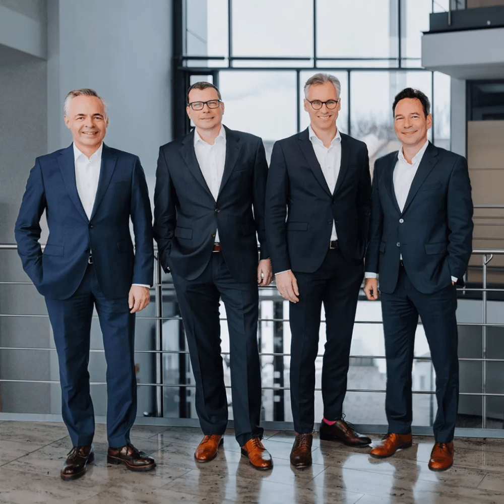 Vier Männer in Anzügen stehen nebeneinander in einem modernen Innenraum mit großen Fenstern im Hintergrund.