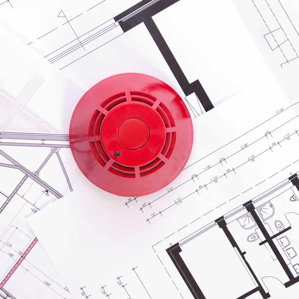 Red fire alarm device placed on architectural floor plans with technical drawings and measurements visible.