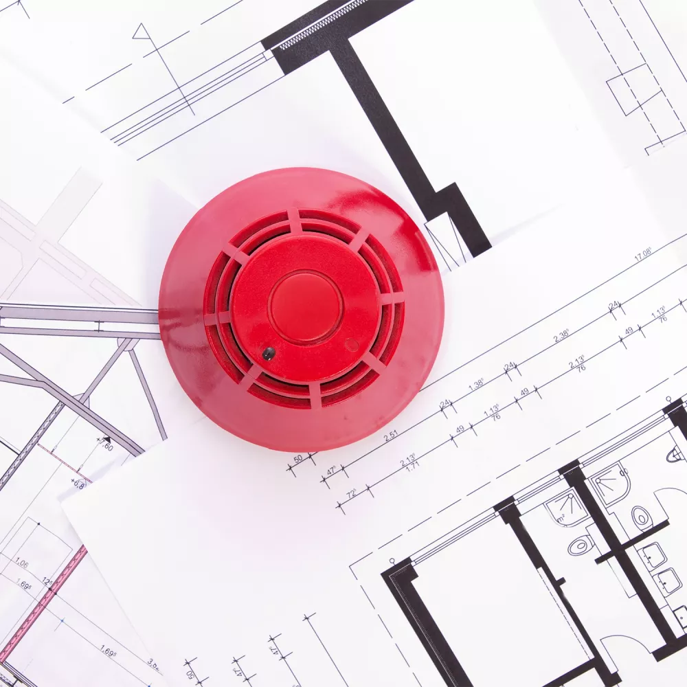 Red fire alarm device placed on architectural floor plans with technical drawings and measurements visible.