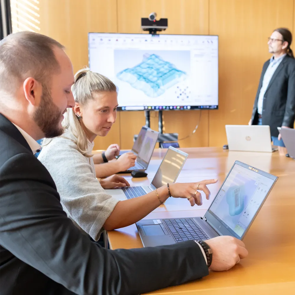Three people working on laptops in a meeting room while a presenter stands near a large screen displaying a 3D model.