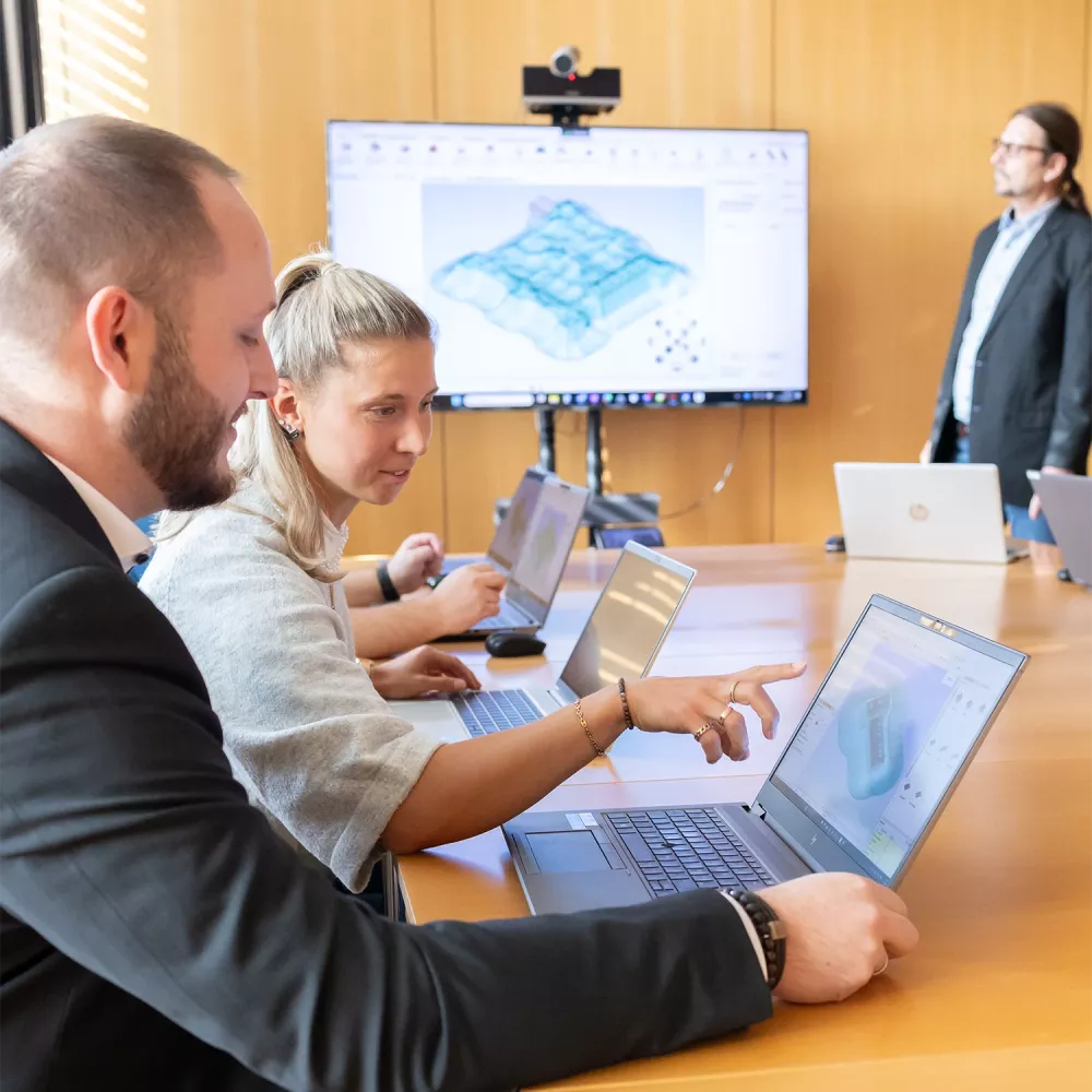 Three people working on laptops in a meeting room while a presenter stands near a large screen displaying a 3D model.