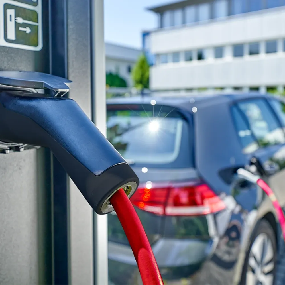 Close-up of an electric vehicle charging connector with red cable plugged into a charging station, car in background.