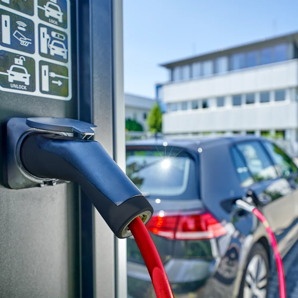 Close-up of an electric vehicle charging connector with red cable plugged into a charging station, car in background.