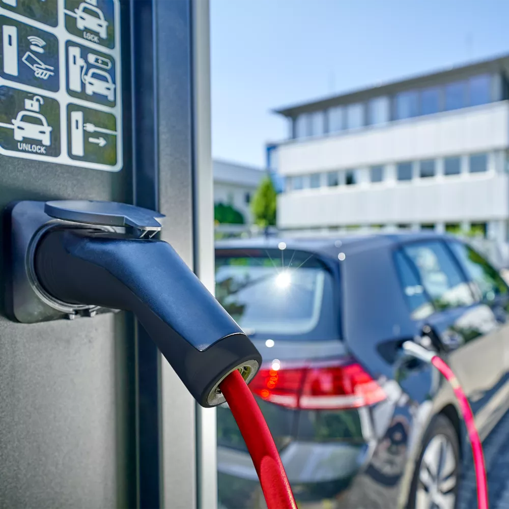 Close-up of an electric vehicle charging connector with red cable plugged into a charging station, car in background.