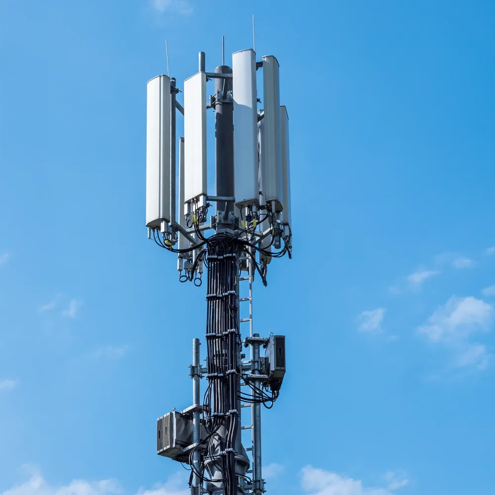 Cellular base station tower with multiple antenna panels and cables against clear blue sky.