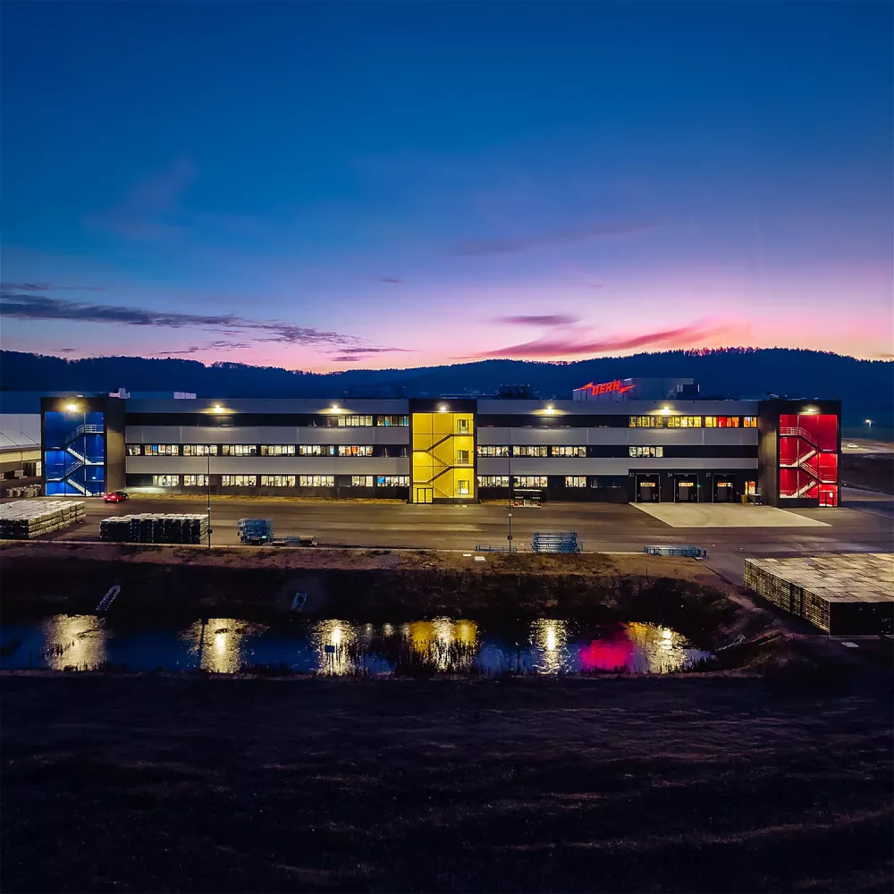 Illuminated industrial building at dusk with lit windows, colored stairwells, a reflective water area in front, and a twilight sky.