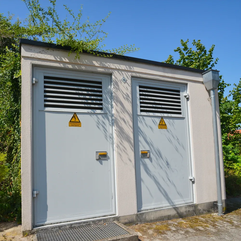 Small gray building with two doors each having ventilation grilles and high voltage warning signs, surrounded by trees.