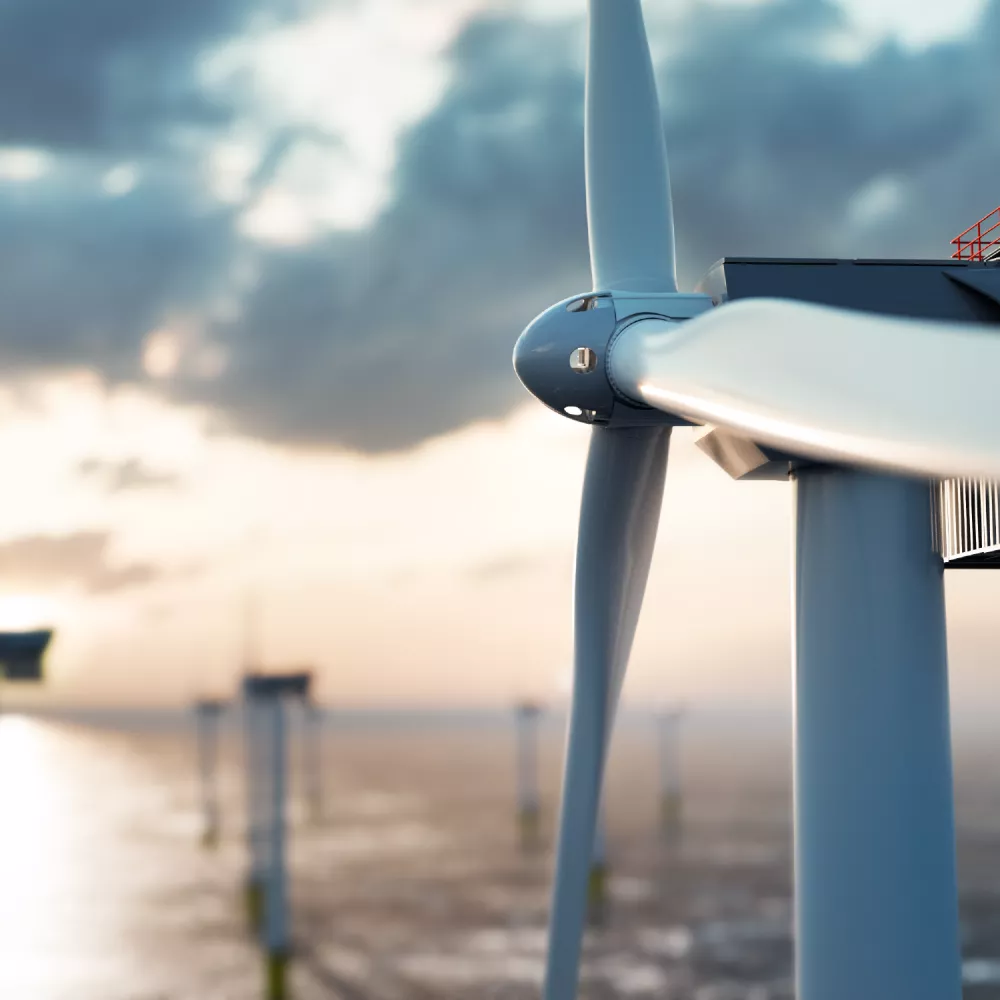 Close-up of a wind turbine blade and hub with other turbines visible in the background over water under a cloudy sky.