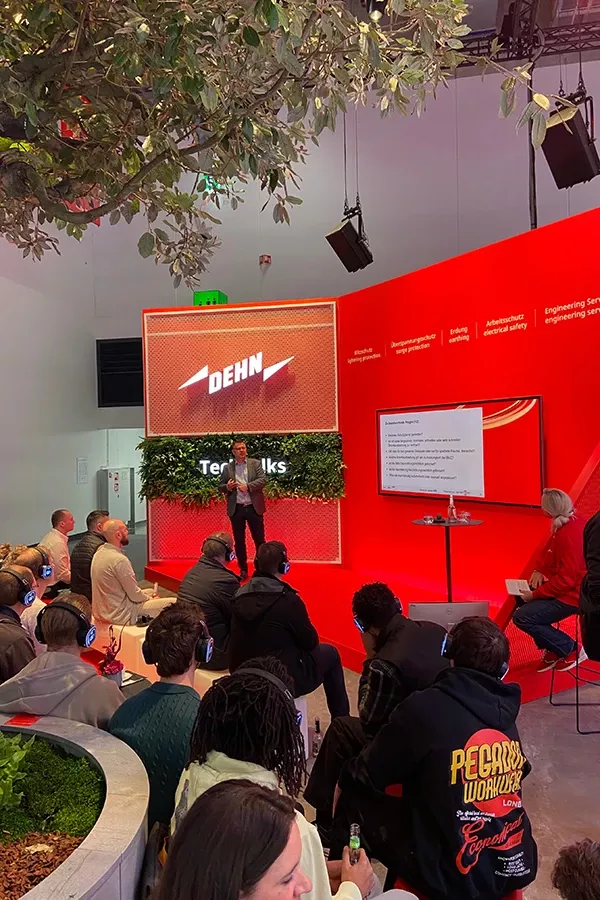 A speaker presents at a DEHN event in front of a seated audience, with a large screen displaying slides and red walls featuring company branding.