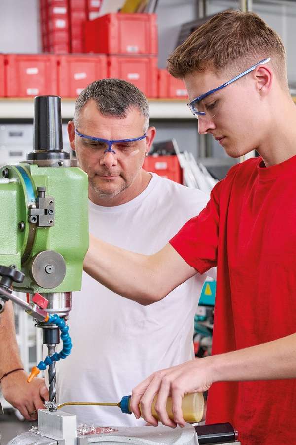 Zwei Männer mit Schutzbrillen arbeiten in einer Werkstatt an einer grünen Bohrmaschine. Der eine bedient die Maschine, der andere beaufsichtigt ihn und beobachtet ihn dabei aufmerksam.
