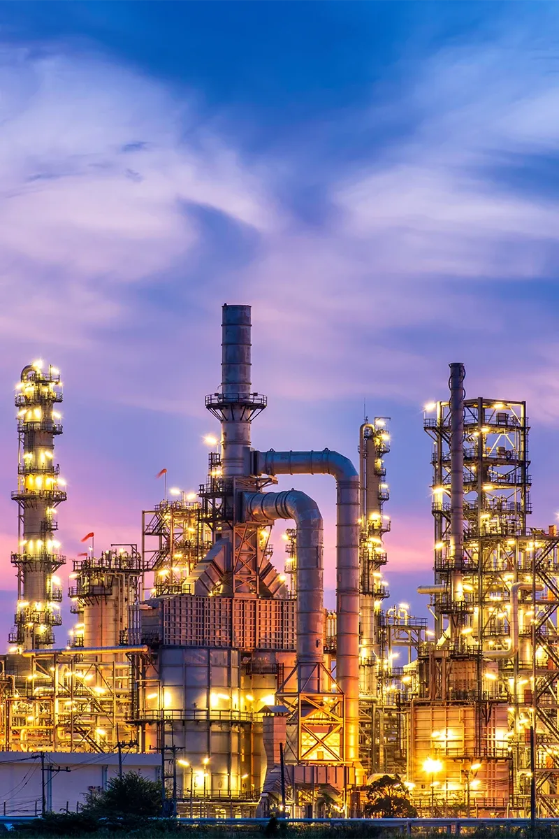 Beleuchtete Chemieanlage im Abendlicht mit zahlreichen Türmen, Pipelines und Lichtern vor einem lila gefärbten Himmel. 