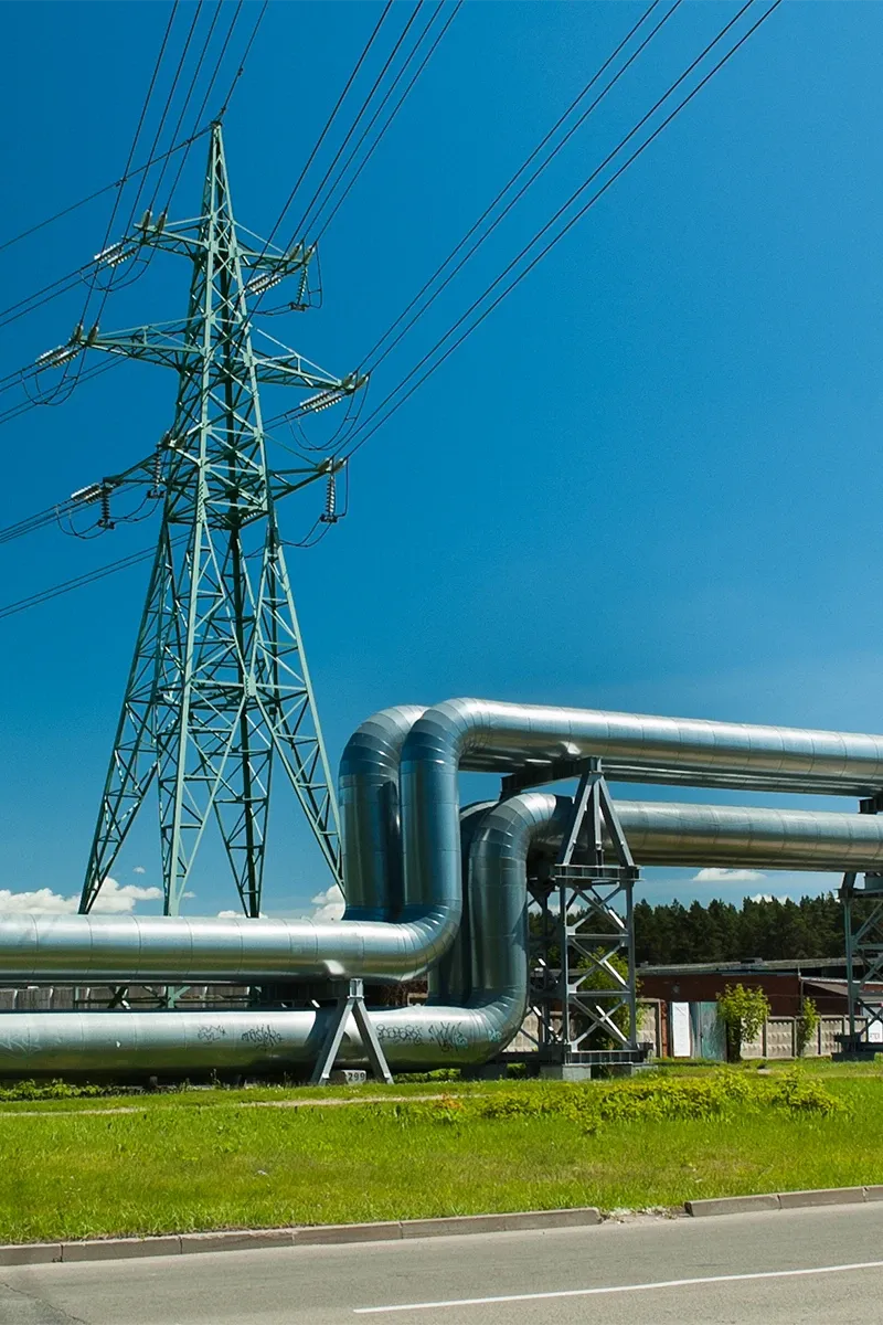 Großer grüner Strommast hinter einer Pipeline auf einer Wiese unter blauem Himmel. 