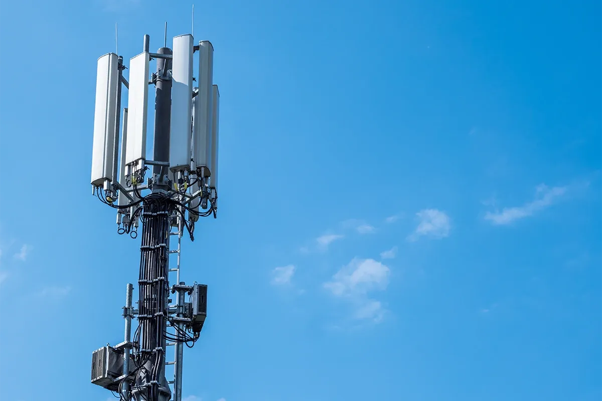 Cell tower with multiple rectangular antennas, cables, and equipment boxes mounted on a mast against a blue sky.