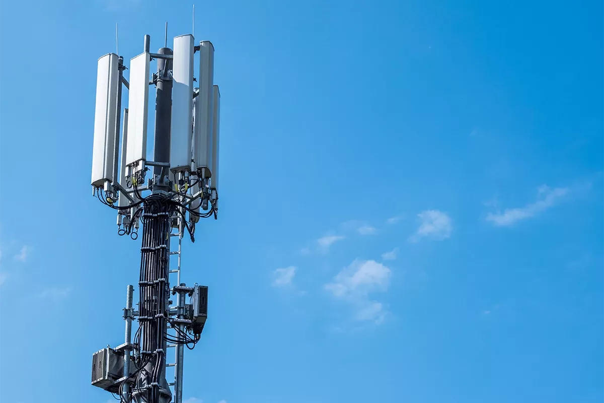 Cell tower with multiple rectangular antennas, cables, and equipment boxes mounted on a mast against a blue sky.