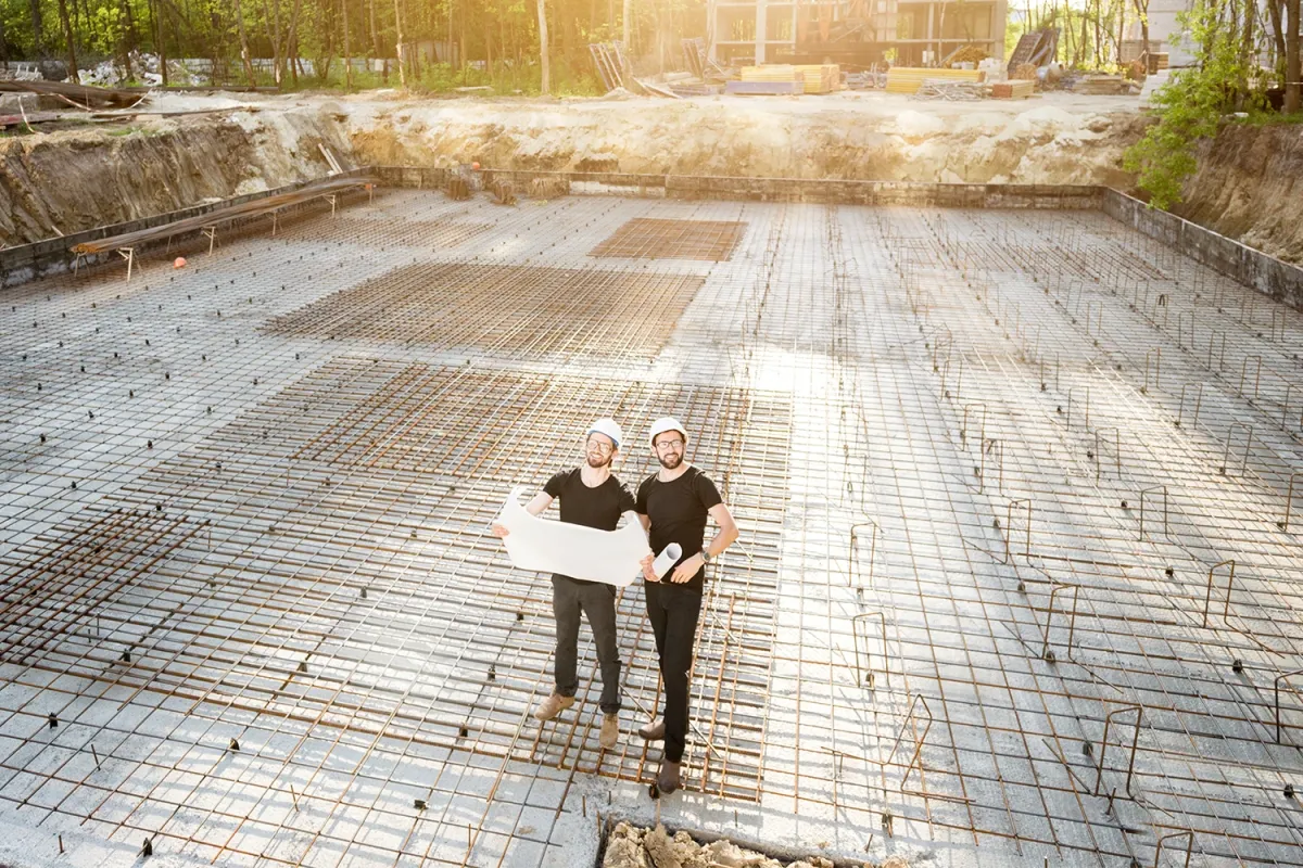 Two construction workers wearing hard hats stand on a reinforced concrete foundation slab, holding blueprints at a building site.