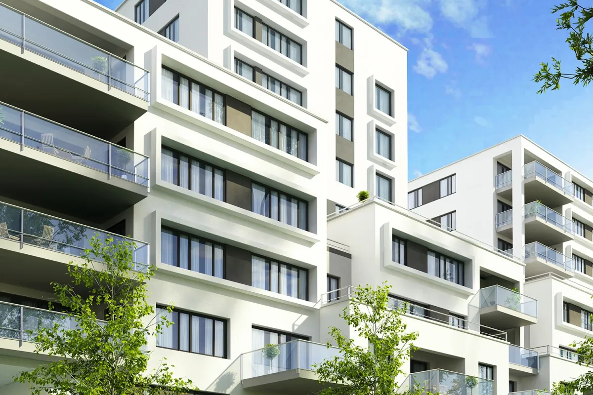 Modern white apartment buildings with multiple balconies, large windows, and surrounding green trees under a clear blue sky.