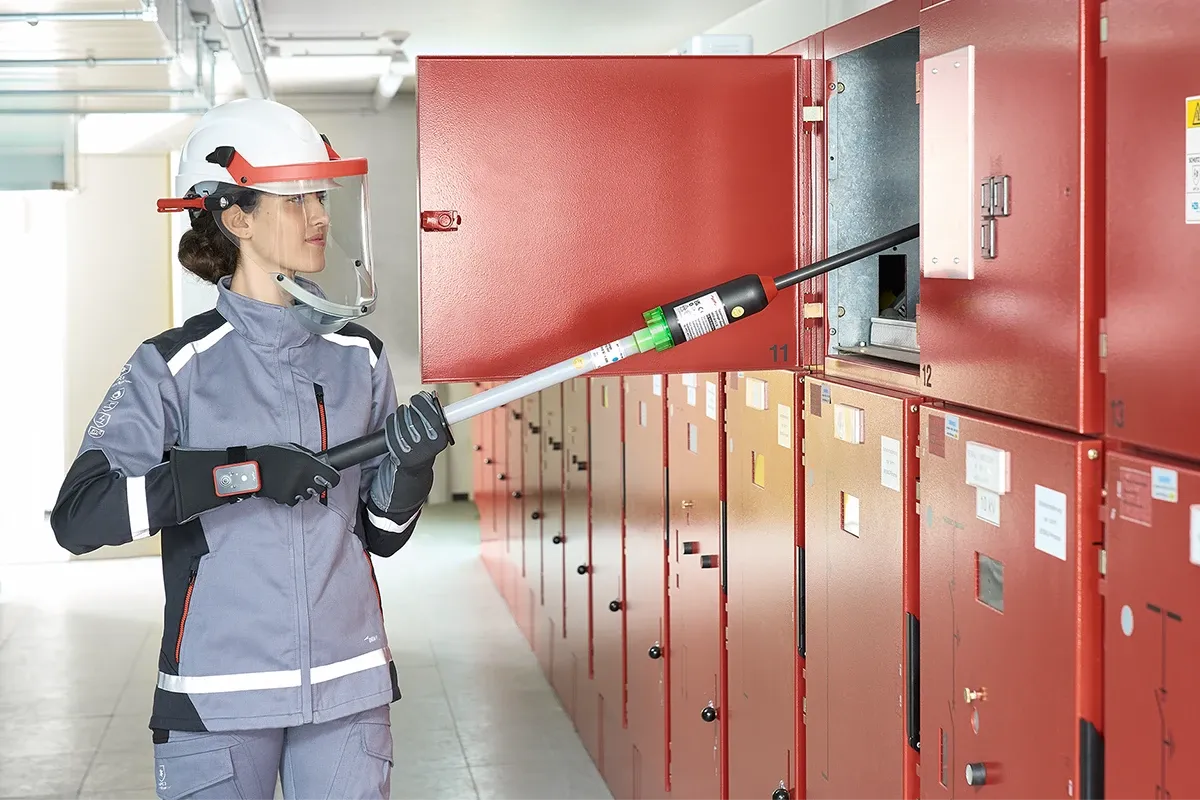 Technician wearing face shield and protective clothing uses an insulated testing pole on an open red electrical switchgear cabinet.