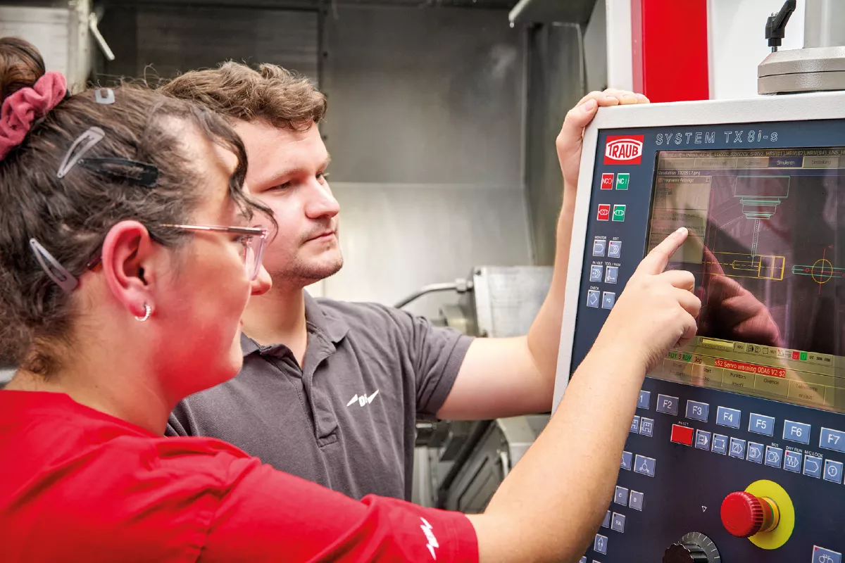 Two people operate a machining control panel with a screen showing technical diagrams.