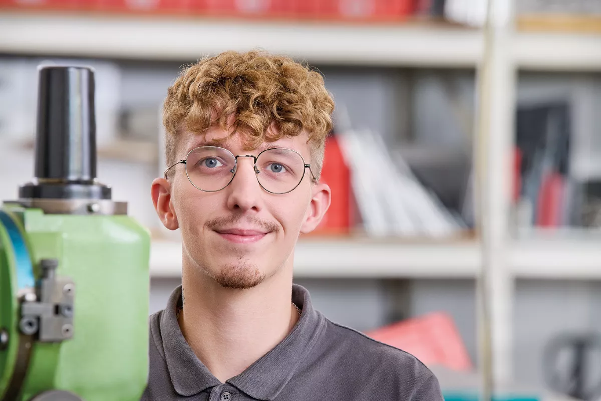 Young man with curly hair wearing a dark polo shirt standing in a workshop with machinery and shelves in the background.