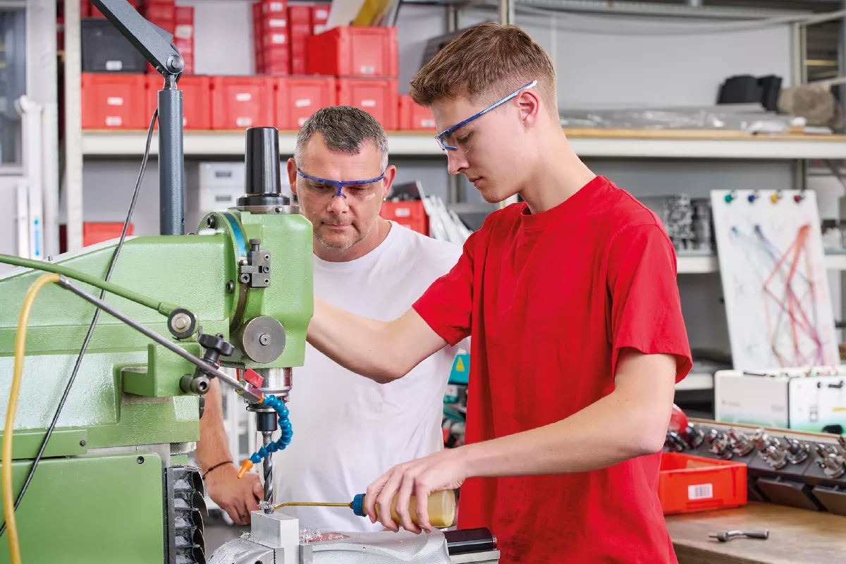 Two young men operating a green industrial machine in a workshop with shelves and red storage bins in the background.