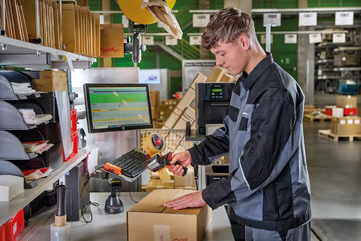 Person scanning a barcode on a cardboard box in a warehouse with shelves and computer equipment.